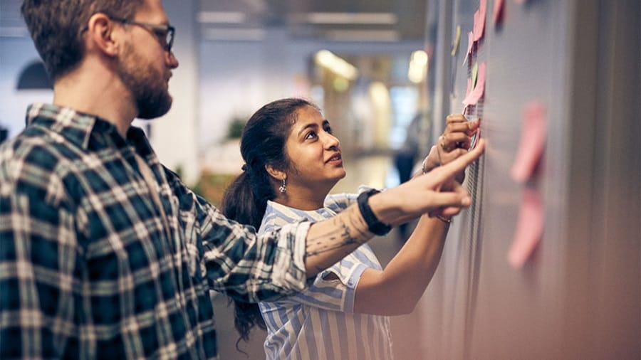 Two individuals in a work environment, interacting with sticky notes on a glass wall, indicating a planning or brainstorming session.